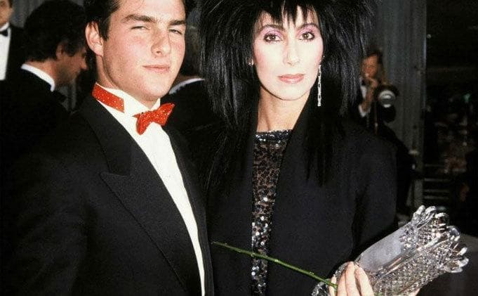 Cher appears holding a rose and a vase and Tom Cruise’s arm. 