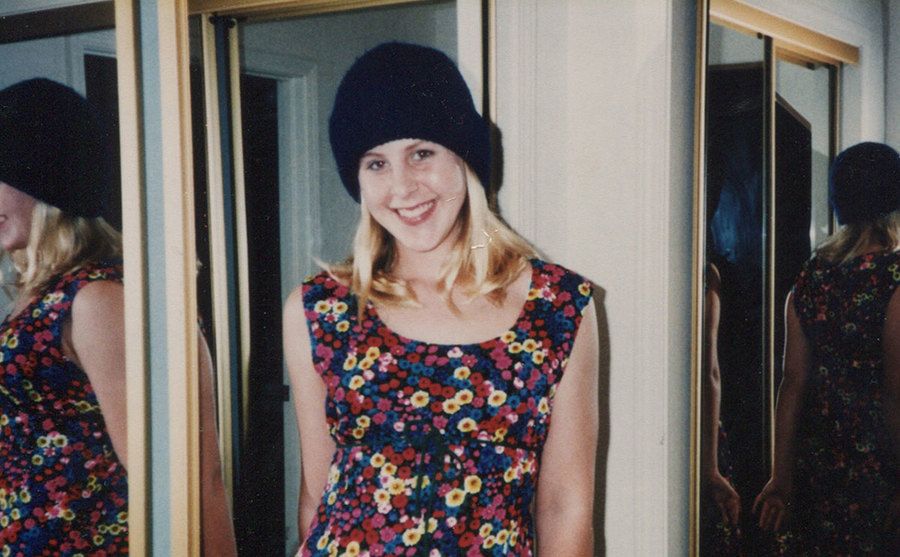 Elyse Pahler smiling next to a mirror wearing a dress and a hat. 