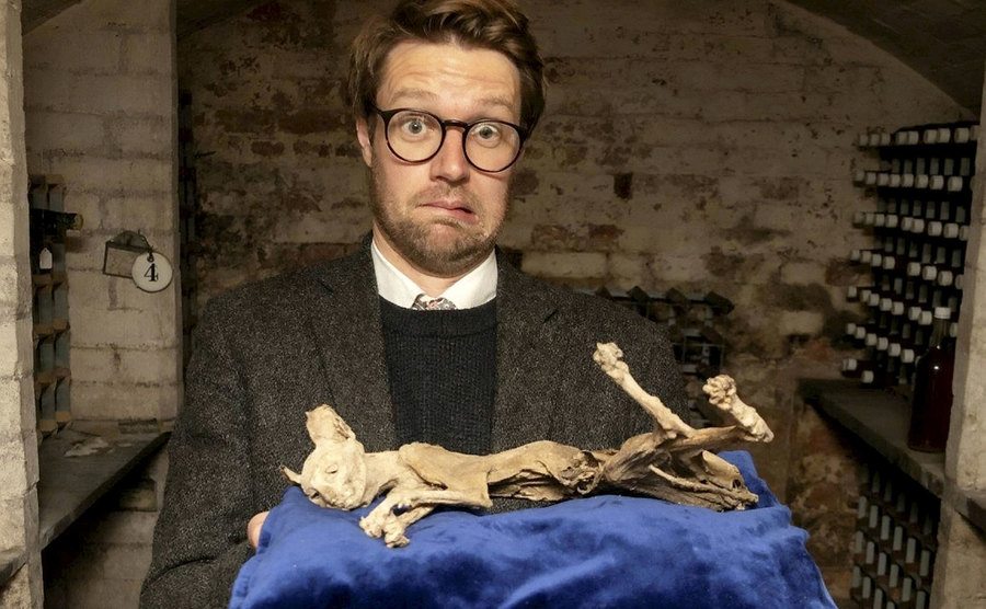 A confused-looking man holds up the mummified cat. 