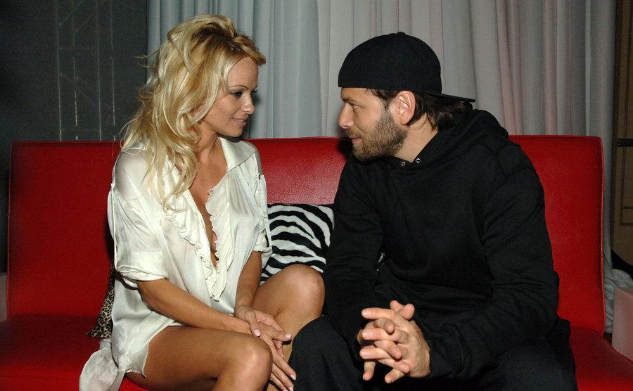 Pamela Anderson e Rick Salomon sentados num sofá vermelho numa festa conversando