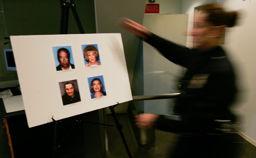 An officer in court holds a poster containing pictures of the defendants and their victims.